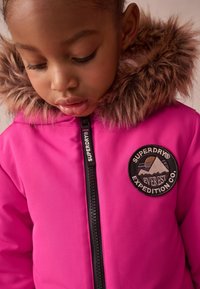 Fuksijas rozā jaka ar mākslīgā kažokāda apdari uz kapuces, ar rāvējslēdzēja priekšpusi un apaļu melnu plāksteri, kurā redzams "Superdry" un kalnu grafikas detaļa.