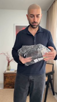 Uomo con barba curata e testa calva che tiene un pacco avvolto in plastica nera in una stanza luminosa con mobili e tende.