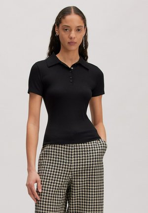 Vrouw die een nauwsluitende zwarte short-sleeve polo en zwart-wit geruite broek draagt, staand met één hand in de zak.