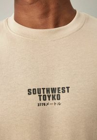Beige puuvillainen crewneck-t-paita, jossa on musta graafinen painatus, jossa lukee "SOUTHWEST TOYKO" ja "3776メートル" edessä.