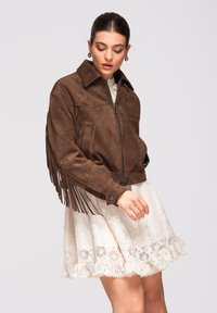 Donna che indossa una giacca con frange in suede marrone sopra un abito bianco di pizzo, guarda verso il basso con una mano leggermente sollevata.