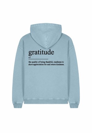 Sweat-shirt à capuche, tissu bleu clair. Le dos présente le mot "gratitude" en grandes lettres noires, avec une définition en dessous en lettres plus petites.