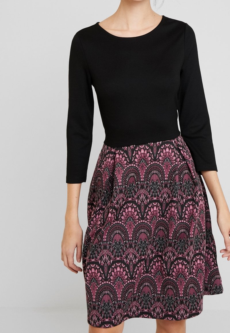 Femme portant une robe longueur genou avec un haut noir uni et une jupe à motifs violets et noirs, debout devant un fond clair uni.