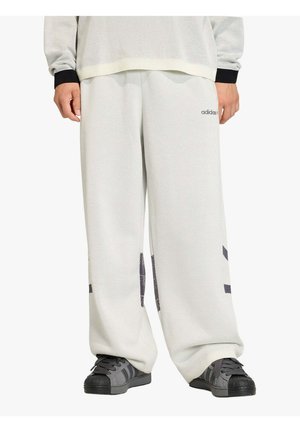 Persona che indossa pantaloni sportivi bianchi Adidas larghi con motivi a blocchi neri e scarpe da ginnastica Adidas nere, in piedi con le mani rilassate ai lati.