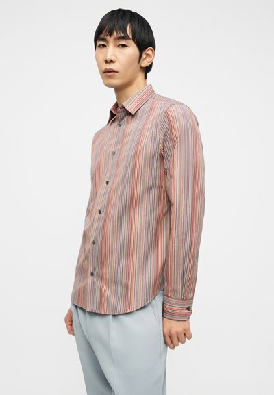 Άνδρας που φοράει πολύχρωμο πουκάμισο με κάθετες ρίγες και ανοιχτό γκρι παντελόνι, στέκεται μπροστά σε απλό λευκό φόντο.