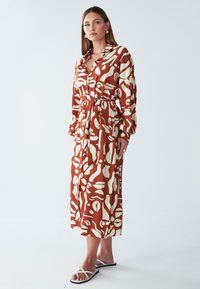 Haljina u boji hrđe i krem s apstraktnim uzorcima, dugim rukavima, prednjim gumbima, pojasom u struku i dužinom do gležnjeva. Glatki materijal.