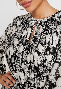 Svartvit blommönstrad klänning med plisserad textur, nyckelhålsknäppning och långa ärmar. Har knäppning med knappar och cinchad midja.