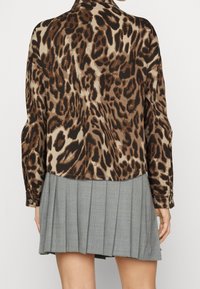 Leopard print shirt in shades of brown and beige, featuring long sleeves and a rounded hem, paired with a pleated gray skirt.