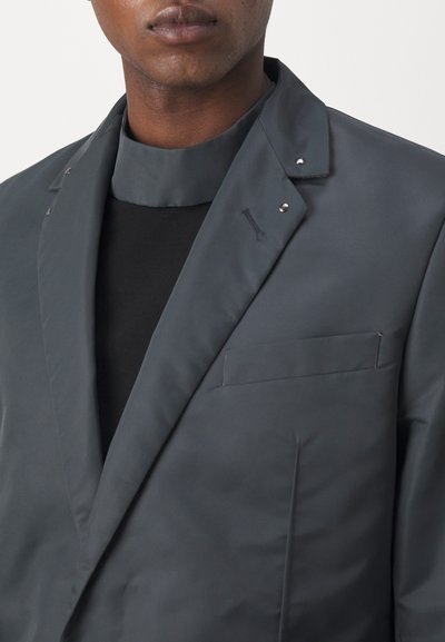 Dunkelgrauer Blazer mit strukturiertem Kragen, auffälliger Nahtführung und einer einzelnen Brusttasche. Glatte, matte Textur mit dezenten Hardware-Akzenten.