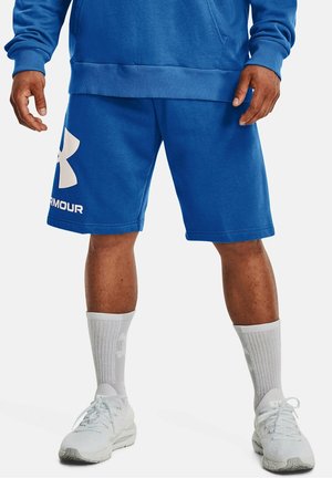 Hombre vistiendo pantalones cortos azules de Under Armour, sudadera azul a juego, calcetines altos blancos y zapatillas deportivas blancas de pie sobre un fondo blanco.