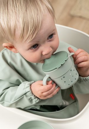 Roheline laste kruus ümarate kaanega, millel on täpiline muster. Valmistatud plastikust, mõeldud väikestele kätele, seda hoiab rohelist jope kandev laps.