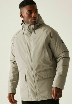 Mężczyzna w beżowej zimowej kurtce z kapturem i przednimi kieszeniami, patrzący na bok na jednolitym jasnym tle.