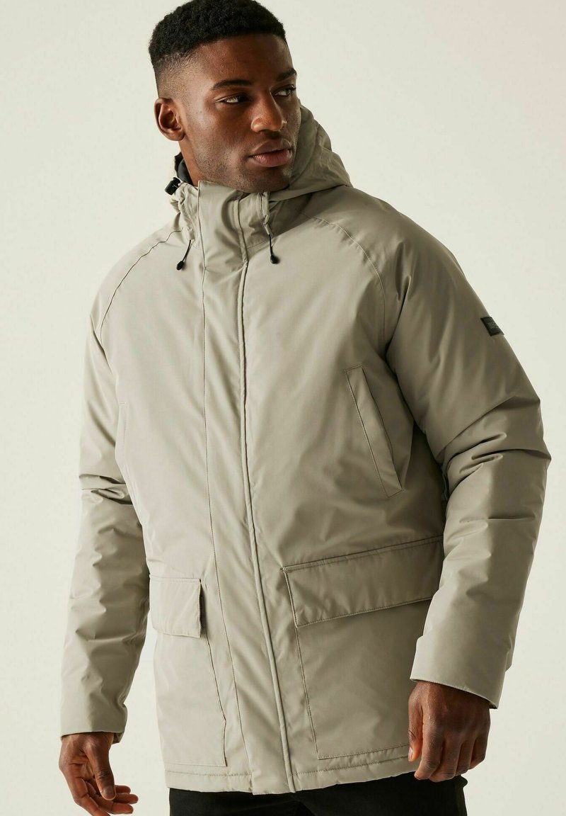 Man wearing beige hooded winter jacket with front pockets, looking sideways against plain light background.