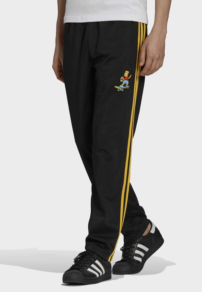 Svarta träningsoverallsbyxor i syntetiskt material, med gula sidostråkar och en färgglad grafik av Bart Simpson på en skateboard på vänster lår.