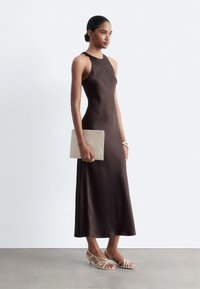 Femme en robe longue sans manches marron foncé tenant une pochette beige tissée, portant des talons blancs à brides, debout devant un fond clair uni.