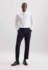 Witte overhemdd met een borstzak en marineblauwe pantalon. Het model draagt zwarte leren schoenen, met de handen in de zakken, tegen een lichte achtergrond.