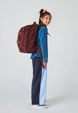 Ragazza giovane che sorride, indossa un grande zaino color marrone scuro, una giacca di jeans e pantaloni bicolore, in piedi davanti a uno sfondo semplice.