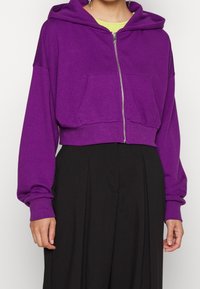 Felpa viola corta con zip frontale, grandi tasche e maniche lunghe, realizzata in un materiale morbido. Indossata sopra una maglietta verde chiaro.