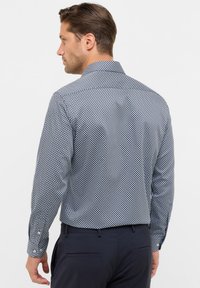 Man in een overhemd met lange mouwen en een kleine diamanten patroon, gecombineerd met donkere broek, met zijn rug naar de camera gekeerd.