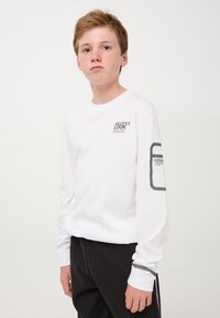 Maglietta bianca a maniche lunghe con grafica "NEVER LOOK BACK" sul petto, dettagli neri sulle maniche, abbinata a pantaloni della tuta scuri.