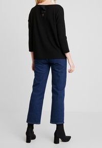 Schwarzes, langärmliges Oberteil mit Rundrücken und einer kleinen Schleifendetail. Getragen mit hoch taillierten blauen Jeans und schwarzen, hochhackigen Stiefeletten.