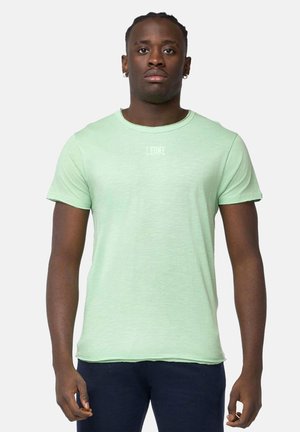 Uomo con capelli corti intrecciati che indossa una maglietta verde chiaro a maniche corte con la scritta "LEONE", in piedi contro uno sfondo bianco semplice.