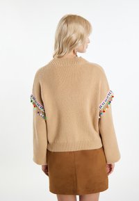 Maglione beige lavorato a maglia con colletto a coste e frange colorate decorative lungo le maniche. Abbinato a una gonna in suede marrone.