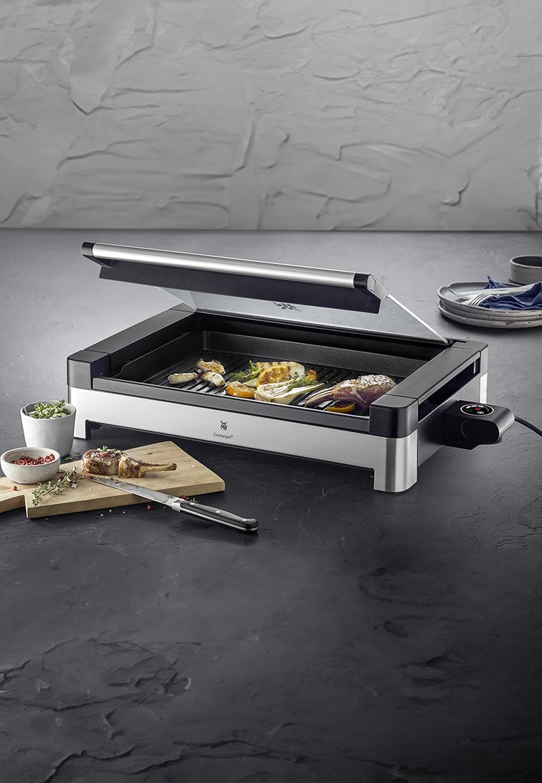 Elektrogrill mit einem Edelstahlrand, schwarzem Gehäuse, transparenter Abdeckung, Grillrillen und einer Auswahl an gegrilltem Gemüse und Fleisch.