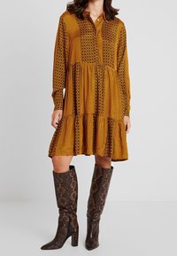 Langärmliges, senfgelbkleid mit gestreiften und floralem Muster, gestuftem Rock. Kombiniert mit kniehohen, braunen, Schlangenlederstiefeln mit Reißverschluss.