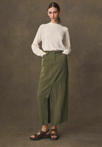 Falda de lino verde oliva con una abertura frontal, cintura alta y bolsillos laterales, combinada con un suéter de punto crema y sandalias marrones.