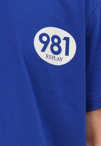 T-shirt in cotone blu con una grafica ovale bianca che presenta i numeri "981" e la parola "REPLAY". Tessuto morbido, maniche corte.