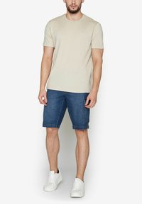 T-shirt beige chiaro con maniche corte, abbinato a pantaloni corti in denim blu scuro con tasca laterale. Sneakers bianche completano l'outfit.