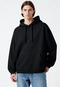 Čierna oversized mikina vyrobená z jemného materiálu, so prednou kapsou, šnúrkou v kapucni a rebrovanými manžetami, v kombinácii so svetlomodrými džínsami.