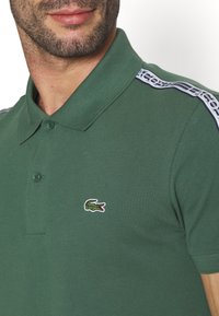 Man wearing green polo shirt with crocodile logo on chest and branded tape on shoulder.