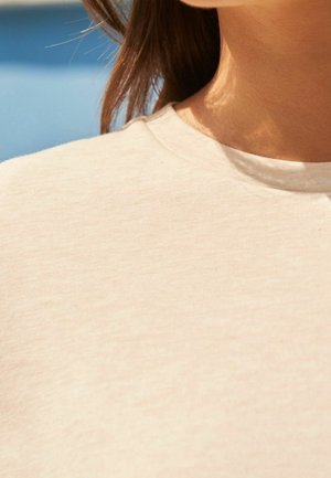 Gros plan sur le cou et l'épaule d'une personne portant un t-shirt beige à col rond, avec des cheveux bruns partiellement visibles sur un fond bleu.