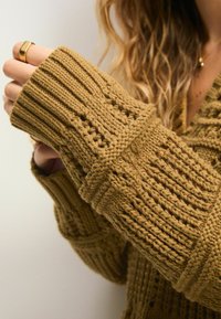 Sennepsfarvet strikket sweater med ribbede og teksturerede mønstre, oversized ærmer og løs pasform. Har en dyb V-halsudskæring og detaljeret syning.