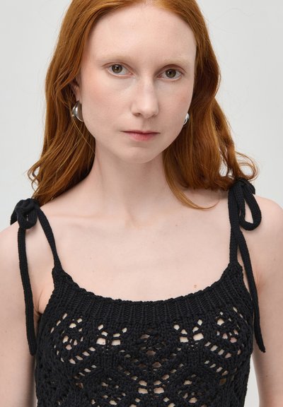 Junge Frau mit roten Haaren trägt silberne Creolen und ein schwarzes gehäkeltes Top mit gebundenen Trägern an den Schultern, blickt geradeaus.