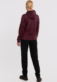 Haut en polaire bordeaux avec un design ajusté, capuche et accents noirs, associé à un pantalon noir fuselé et des chaussures noires robustes.