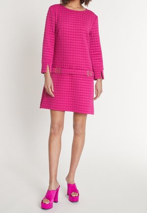 Fuchsia gewatteerde trui en bijpassende rok met een boxy ontwerp, lange mouwen en decoratieve metalen accenten op de taille. Roze plateauhakken.