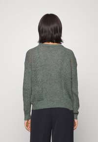 Person med kort mörkt hår som bär en texturerad grön stickad tröja och svarta byxor, vänd bort från kameran mot en enfärgad ljus bakgrund.