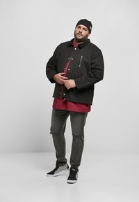 Giacca nera in tela con bottoni a pressione, tasche frontali e una zip argentata; abbinata a una camicia bordeaux, jeans scuri strappati e sneakers in bianco e nero a quadri.