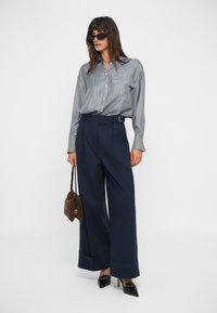 Chemise à rayures bleu et blanc avec des boutons, associée à un pantalon large marin avec une ceinture et des ourlets à revers. Les accessoires comprennent un sac à main marron.