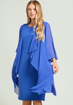 Blonde Frau in einem knieumspielenden königsblauen Kleid mit einem passenden, fließenden, transparenten blauen Schal, die eine Seite des Schals hält.