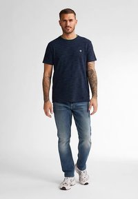 Tricou cu dungi bleumarin, cu guler verde, mâneci scurte și un logo mic pe piept, asortat cu blugi albastru deschis și adidași albi.