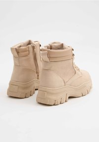 Par de botas de tobillo de gamuza beige con suelas de goma gruesas, cordones en la parte delantera y cremalleras en los laterales, exhibidas sobre un fondo blanco.