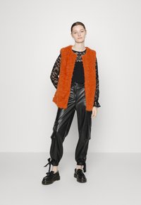 Colete de pele falsa laranja sobre uma blusa preta de renda de manga longa, combinado com calças cargo de pele de cabra pretas e sapatos pretos de atacador.