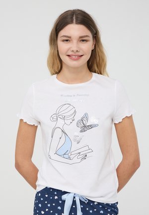 Giovane donna che sorride, indossando una maglietta bianca con una linea che rappresenta una donna che tiene in mano libri e farfalle, abbinata a pantaloni blu con motivi a cuore.