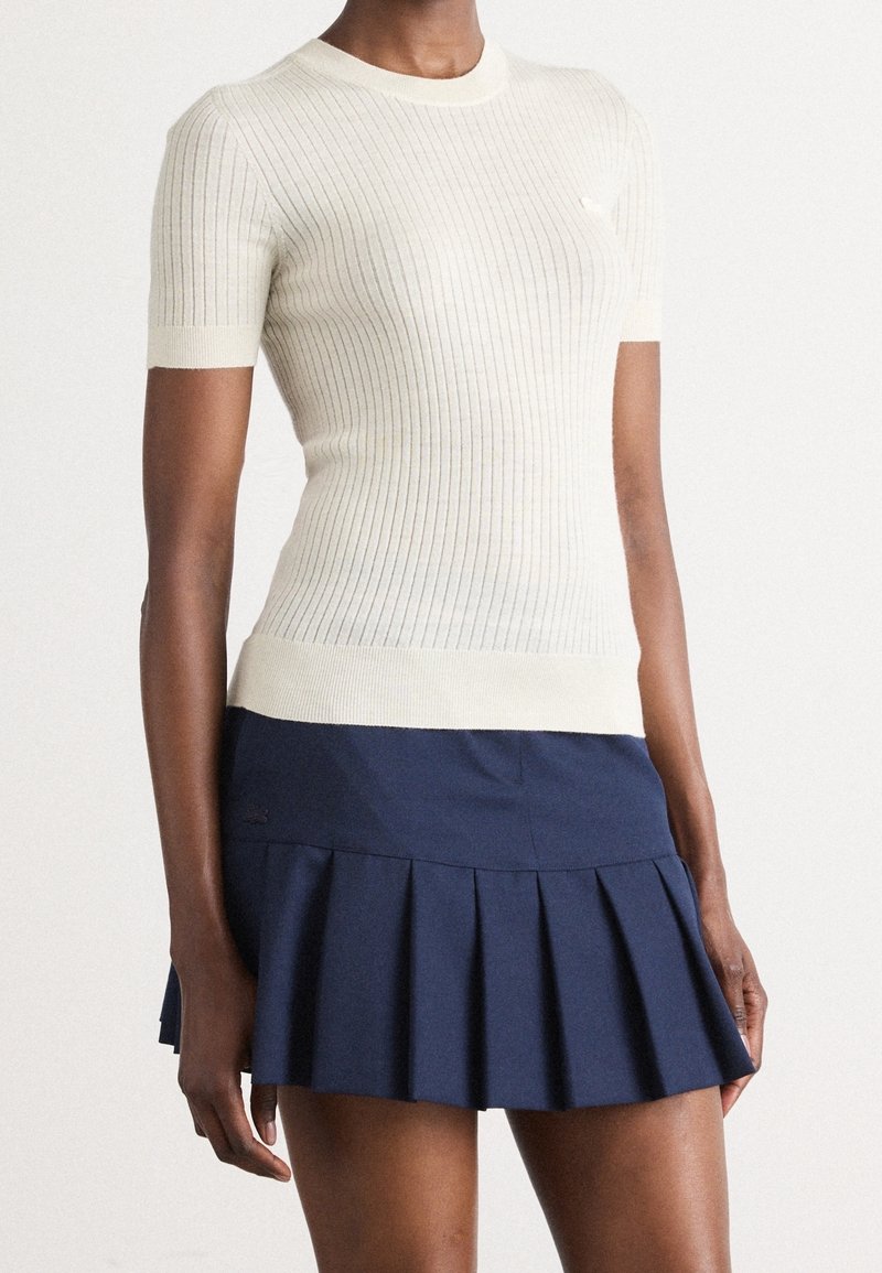 Pull crème à manches courtes côtelé et ajusté, associé à une mini-jupe plissée bleu marine sur une personne debout.