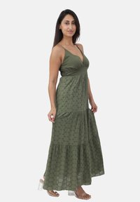 Abito lungo verde oliva con corpino aderente e gonna a strati e текстurizada caratterizzata da motivi circolari e spalline sottili. Tacchi a blocco trasparenti.