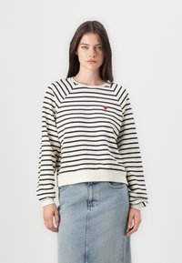 Stripet genser i krem og svart, med et rødt hjerteemblem, rund hals og ribbede mansjetter. Kombinert med en denimskjørt.